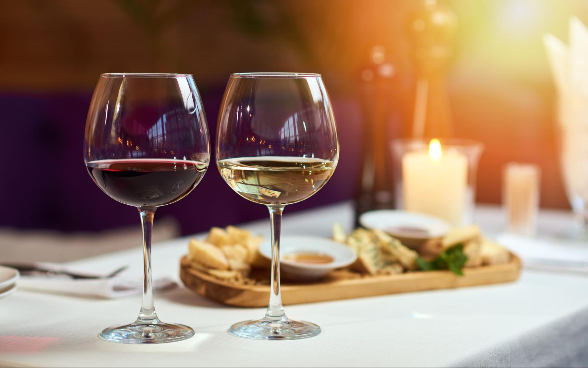 Two wine glasses on a table, one with red wine and one with white wine. There are snacks and candles on the table.
©Kartashov Stas