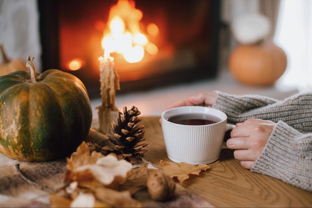 Hands holding a cup of tea on a wooden table with a small pumpkin and autumn leaves. The glow of a warm fireplace is in the background.
©Bogdan Sonjachnyj