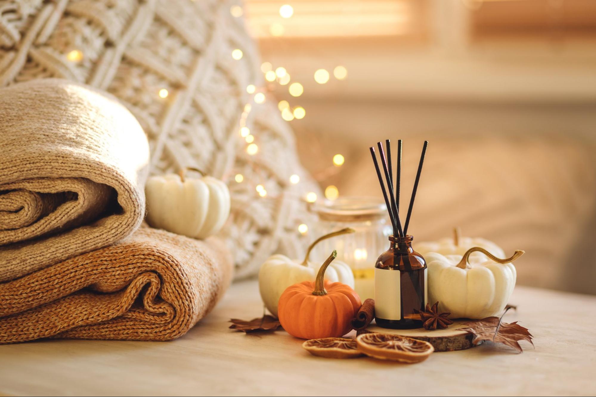 Stacked knit blankets, mini pumpkins, dried leaves, and other fall decor on a table with sparkling lights in the background ©Vera Prokhorova