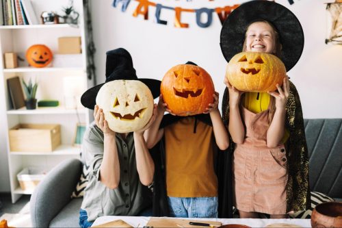 Woman and two children playfully hiding their faces behind carved pumpkins with Halloween decorations in the background ©AYO Production