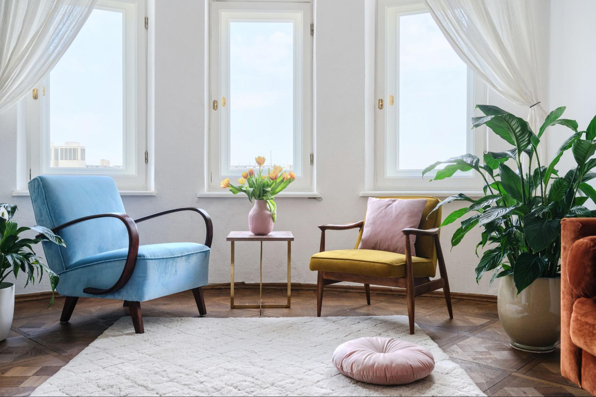 Bright living room with sheer curtains, blue armchairs, tulips in a pink vase, and indoor plants. ©brizmaker