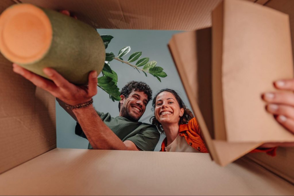 A happy couple smiling while looking into a moving box and unpacking a plant. ©Zamrznuti tonovi