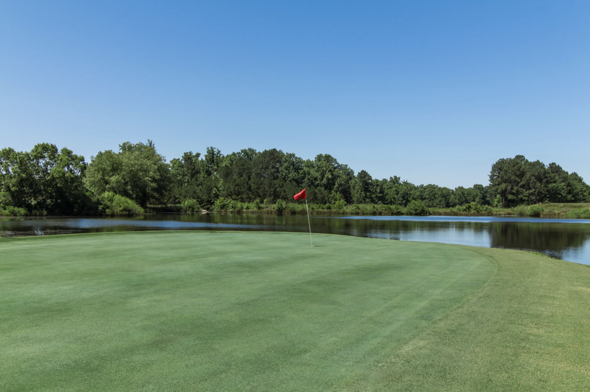 A pristine green on the Traditions of Braselton golf course featuring a red flag next to a scenic pond.