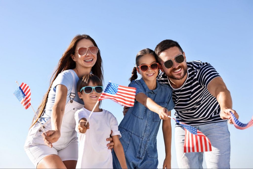 A happy family wearing sunglasses and smiling while waving small American flags under a clear blue sky. ©New Africa