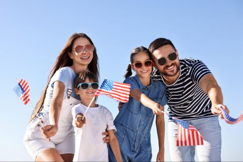 A happy family wearing sunglasses and smiling while waving small American flags under a clear blue sky. ©New Africa