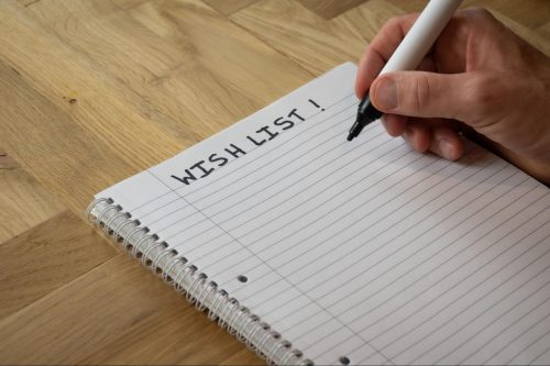 Person writing a home project wishlist on a notepad sitting on a wooden table. ©Drew McArthur
